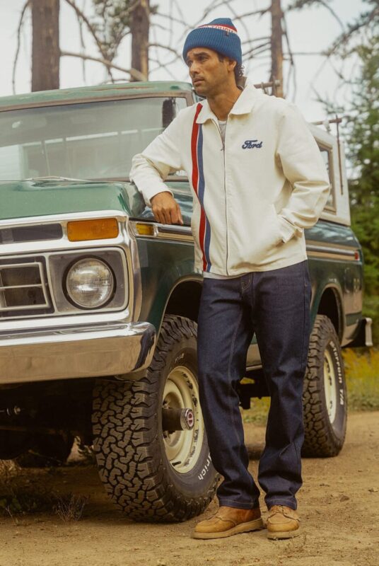 A man in a white Ford x Brixton jacket, jeans, tan shoes, and a blue beanie leans against an older green pickup truck outdoors near trees.