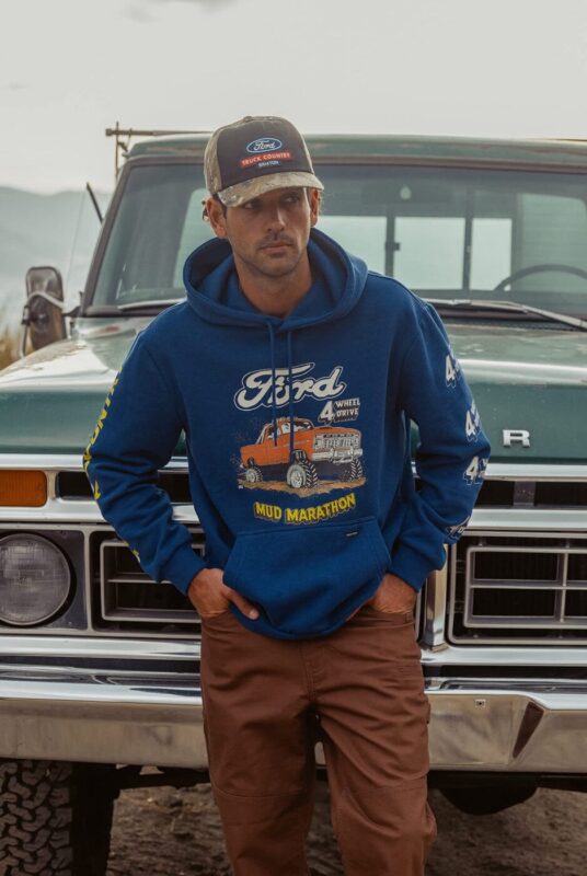 A man wearing a blue Ford x Brixton hoodie, brown pants, and a Ford cap stands in front of a vintage green Ford truck from the Limited Edition Collection.