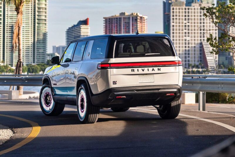 A white Rivian SUV, inspired by the Rivian Opens Brickell Hub with a One-Off Miami-Themed Tri-Motor R1S, cruises along a curved urban road with sleek high-rise buildings in the background.