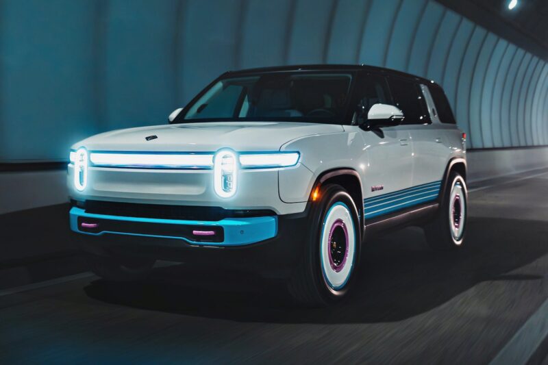 A white electric SUV, reminiscent of Rivian’s one-off Miami-themed tri-motor R1S, cruises through a modern tunnel with illuminated headlights and blue accent lighting.