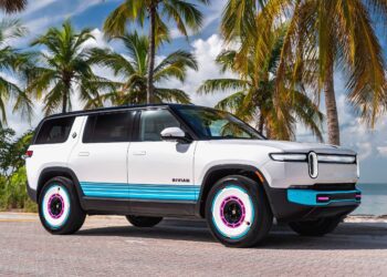 A white Rivian SUV with blue and pink accents is parked on a palm-lined road by the beach—echoing the vibe of the Rivian Opens Brickell Hub With a One-Off Miami-Themed Tri-Motor R1S under a partly cloudy sky.
