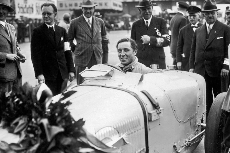 A smiling race car driver sits in his vintage car surrounded by men in suits and hats at what appears to be a racing event, capturing an era that Bugatti honors with the Veyron’s 20-year legacy and special logo.