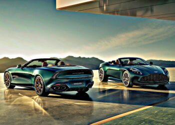 Two green Aston Martin Vanquish convertibles, part of the Q by Aston Martin Marks 60 Years of Volante with Ultra-Exclusive Anniversary Editions, are parked on a sleek terrace with mountains behind and sunlight glinting off their curves.