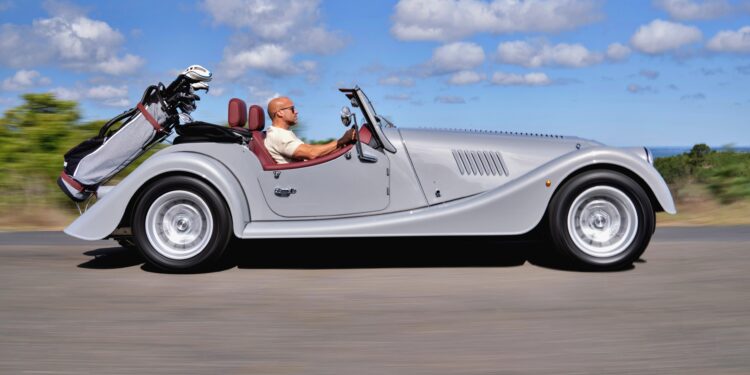 A man drives a silver Morgan Plus Four convertible with a set of golf clubs in the back, cruising down an open Long Island road under a partly cloudy sky.