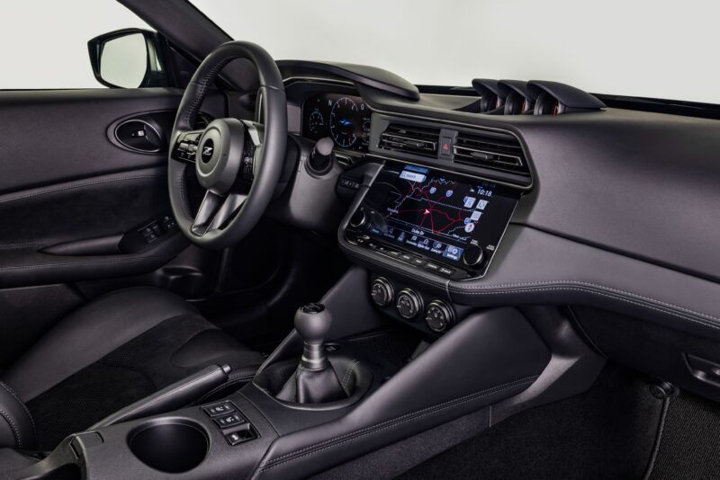 Interior view of a modern car featuring 300ZX-Inspired Design, with manual transmission, black upholstery, digital navigation display, and analog gauges behind the wheel—an homage to the Nissan Z Heritage Edition.