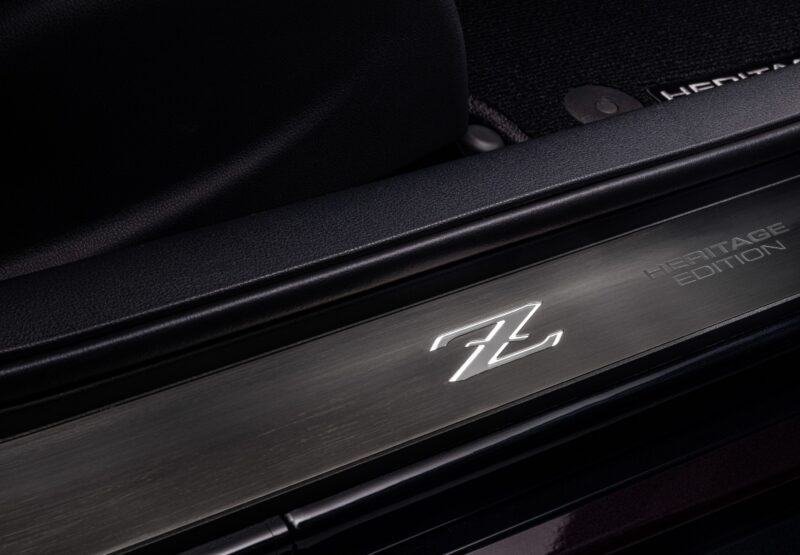 Close-up of an illuminated "Z" logo and "Heritage Edition" text on a metallic car door sill plate, highlighting the 2026 Nissan Z's black interior trim and 300ZX-inspired design.