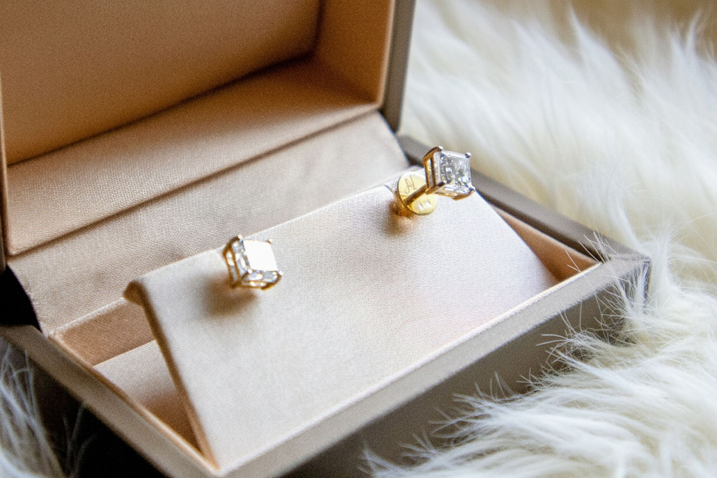 A pair of gold earrings with clear gemstones from AviNika Jewelry is displayed in an open jewelry box on a white fur surface, embodying timeless heirlooms to be cherished for generations.