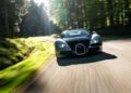A legendary black sports car speeds along a sunlit forest road, with trees and greenery flanking the curved asphalt, its Chassis 5.0 handling each bend with precision.
