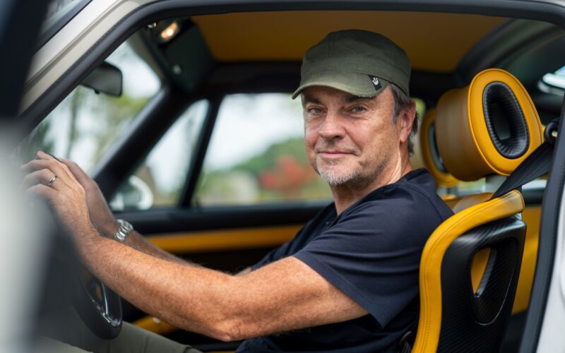 A man wearing a green cap and black shirt sits in the driver's seat of a Porsche 911 Carrera with yellow and black interior, looking at the camera.