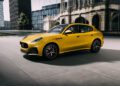 A yellow Maserati Grecale Folgore EV luxury SUV is parked on a modern urban plaza, with glass buildings and reflections in the background.