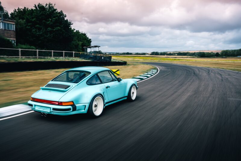 A light blue Porsche 911 by Singer Vehicle Design with white wheels drives on a curved racetrack bordered by grass under a cloudy sky at Fuji Speedway.