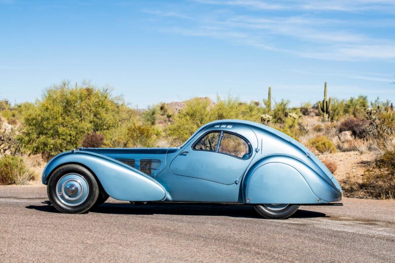 A vintage blue coupe car, echoing automotive history, is parked on a paved road in a desert landscape with shrubs and cacti under a clear sky.