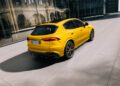 A yellow Maserati Grecale Folgore SUV, featuring advanced EV tech, is parked on a light gray pavement beside a modern glass building on a sunny day.