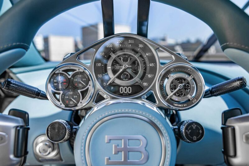 Close-up view of a Bugatti Tourbillon car dashboard, showcasing detailed analog gauges, a digital speedometer display, and the EB logo on the steering wheel—highlighting the brand’s history of precision engineering.