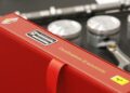 A red binder labeled "Certificazione di autenticità" with Ferrari logos is shown in front of automotive engine parts, including three pistons and a wrench.