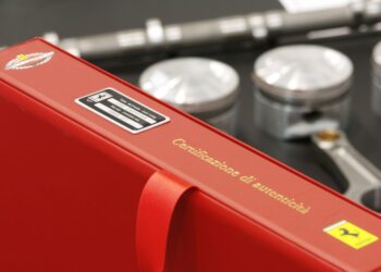 A red binder labeled "Certificazione di autenticità" with Ferrari logos is shown in front of automotive engine parts, including three pistons and a wrench.