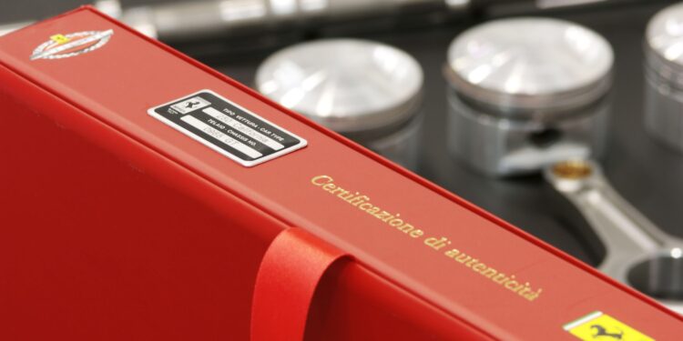 A red binder labeled "Certificazione di autenticità" with Ferrari logos is shown in front of automotive engine parts, including three pistons and a wrench.