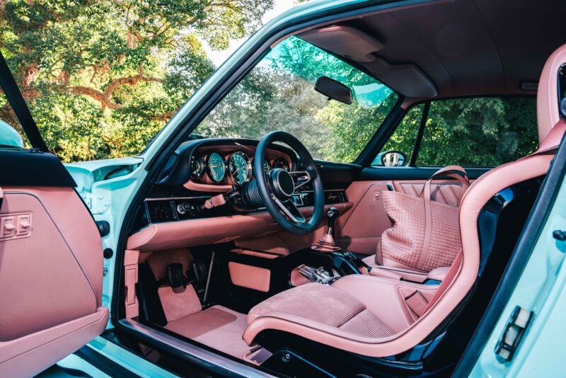 Car interior of a Reimagined Porsche 911 by Singer Vehicle Design, featuring pink leather seats and trim, manual gear shift, steering wheel, and dashboard; driver’s door open with greenery visible outside.