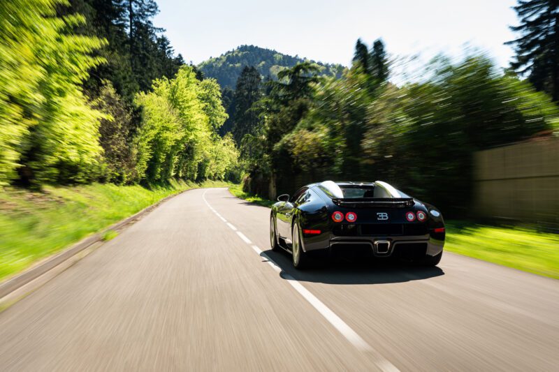 A sleek black Bugatti Veyron sports car speeds along a winding, tree-lined road in a mountainous area on a clear, sunny day.