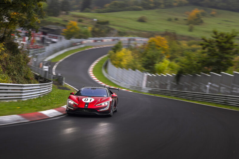 A red electric hypercar marked with number 07 drives around a sharp corner on a racetrack surrounded by greenery and trees, reminiscent of a Nürburgring record attempt.