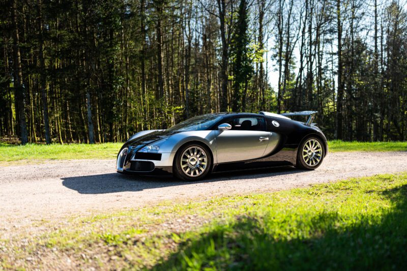 A black and silver Bugatti Veyron sports car is parked on a gravel road with trees and greenery in the background on a sunny day.