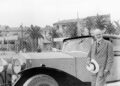 An older man in a suit stands next to a vintage Rolls-Royce, holding a hat, as palm trees and large buildings evoke the timeless elegance of the Phantom, honoring 100 years with a groundbreaking private collection.