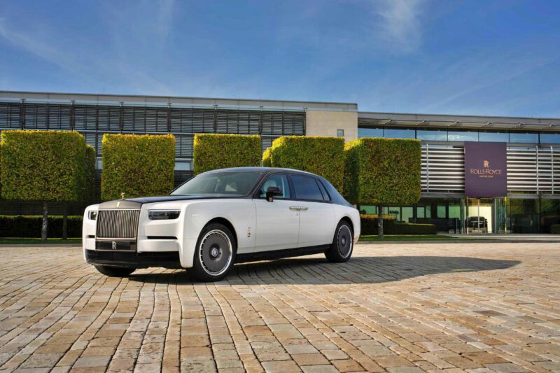 A white Rolls-Royce sedan, part of the Rolls-Royce Honors 100 Years Of Phantom With Groundbreaking Private Collection, is parked on a cobblestone driveway in front of a modern building with neatly trimmed trees and a Rolls-Royce sign.