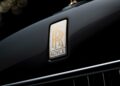 Close-up view of a Rolls-Royce badge on the hood of a black car, showcasing the gold and white "RR" logo with "Rolls Royce" text above a chrome grille as Rolls-Royce honors 100 years of Phantom with groundbreaking private collection.
