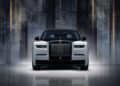 Front view of a white Rolls-Royce Phantom with a black hood, showcased against a dark, elegant background with vertical light streaks, celebrating Rolls-Royce Honors 100 Years Of Phantom With Groundbreaking Private Collection.