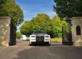A white Rolls-Royce Phantom, part of the Rolls-Royce Honors 100 Years Of Phantom With Groundbreaking Private Collection, glides through black iron gates, framed by stone pillars, with lush green trees and a clear blue sky beyond.