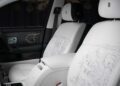 Front seats of a luxury car with white leather upholstery, detailed line art designs, and the Rolls-Royce logo on the headrests celebrate the Rolls-Royce Honors 100 Years Of Phantom With Groundbreaking Private Collection.