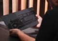A person holds a rectangular metal plate engraved with a vintage car, likely commemorating Rolls-Royce Honors 100 Years Of Phantom With Groundbreaking Private Collection, near a window with vertical wooden slats.