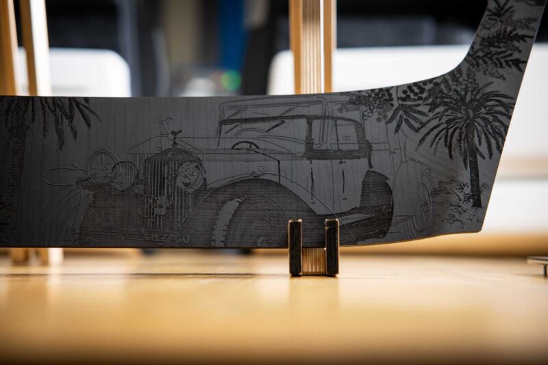 Close-up of a metal sheet engraved with a vintage car scene, featuring two people in the car and palm trees in the background, propped upright—a tribute as Rolls-Royce honors 100 years of Phantom with groundbreaking private collection art.