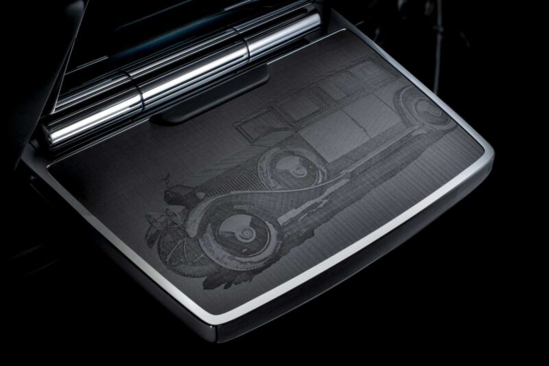 A close-up of a metallic surface engraved with the image of a vintage car, featuring detailed lines and shading, set against a dark background, celebrates Rolls-Royce Honors 100 Years Of Phantom With Groundbreaking Private Collection.