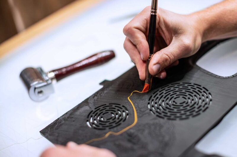 A person uses a tool to trace or cut a pattern on black leather with circular perforations, next to a roller tool—craftsmanship reminiscent of the bespoke attention found in the Rolls-Royce Honors 100 Years Of Phantom With Groundbreaking Private Collection.