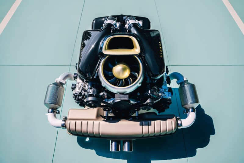 A high-performance Porsche 911 Carrera engine with visible exhaust pipes and air intake is displayed on a light blue surface under bright lighting, embodying the precision of Singer Vehicle Design and the spirit of Fuji Speedway.