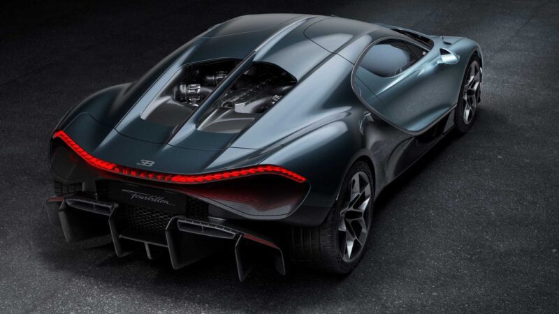 A sleek, dark gray Bugatti hypercar is parked on a textured surface, viewed from the rear, showcasing its aerodynamic design and prominent rear lights—an embodiment of how form, function, past and the future came together in the Bugatti Tourbillon.