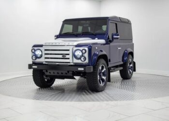 A blue and silver two-door SUV, a unique creation, is parked indoors in a garage on a circular display platform, viewed from the front left angle.