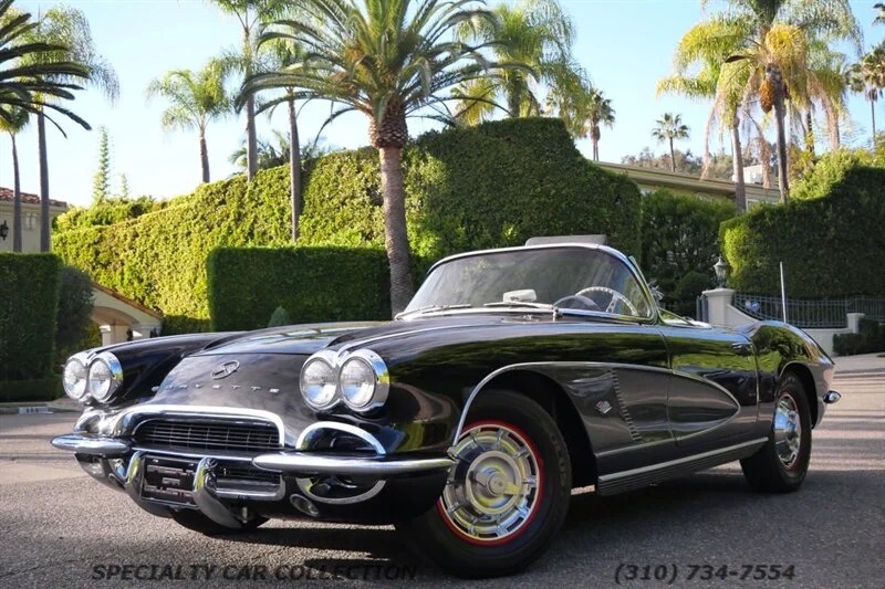 A black vintage convertible sports car, reminiscent of classic muscle cars, is parked on a street lined with palm trees and manicured hedges.