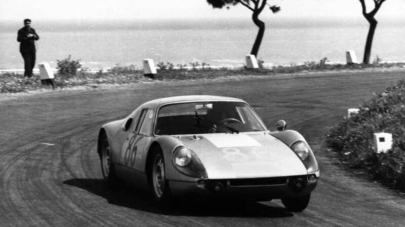 A vintage 904 Restomod race car rounds a curve on a coastal road as a man stands by the roadside in the background; trees and ocean are visible, capturing the spirit of Beyond 911 and Aerfal Automotive craftsmanship.