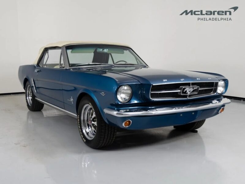 A blue classic Ford Mustang convertible with a beige soft top is parked indoors on a white floor, showcasing iconic Classic Muscle Cars style, with a "McLaren Philadelphia" logo on the wall.