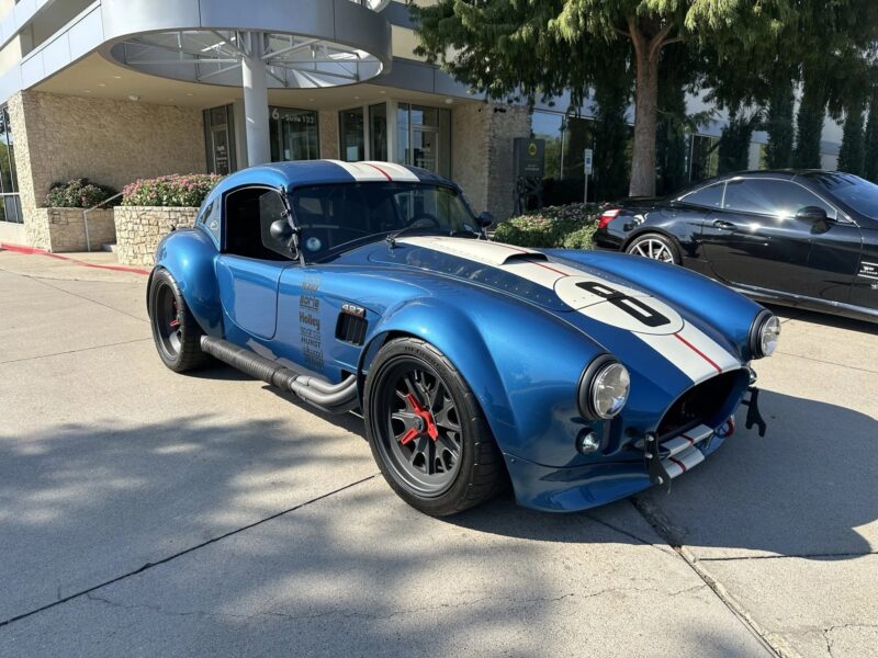 A blue Shelby Cobra classic muscle car with white racing stripes and the number 8 on the hood is parked on a concrete driveway in front of a modern building.