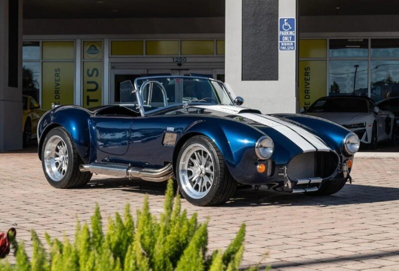 A blue Shelby Cobra with white racing stripes is parked on a brick driveway outside a car dealership, showcasing one of the classic muscle cars for sale, with a "Parking Only" sign visible in the background.