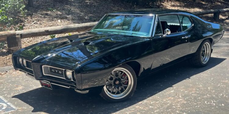 A black classic Pontiac GTO muscle car is parked on a sunlit road near a wooded area, capturing the timeless allure of Classic Muscle Cars with trees and a wooden fence in the background.