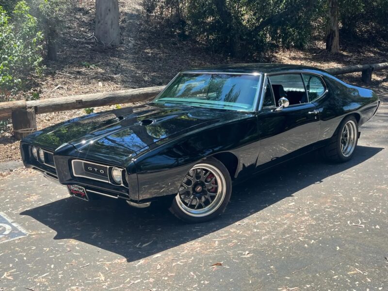 A black classic Pontiac GTO muscle car is parked on a sunlit road near a wooded area, capturing the timeless allure of Classic Muscle Cars with trees and a wooden fence in the background.