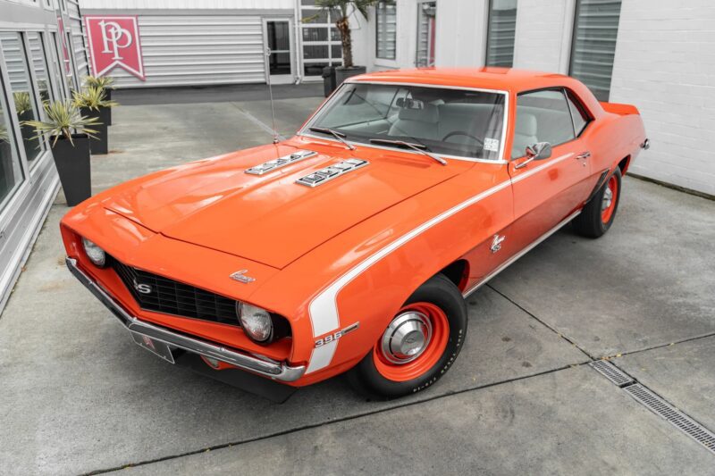 An orange 1969 Chevrolet Camaro SS coupe, a true icon among classic cars, is parked on a concrete driveway next to a white building.