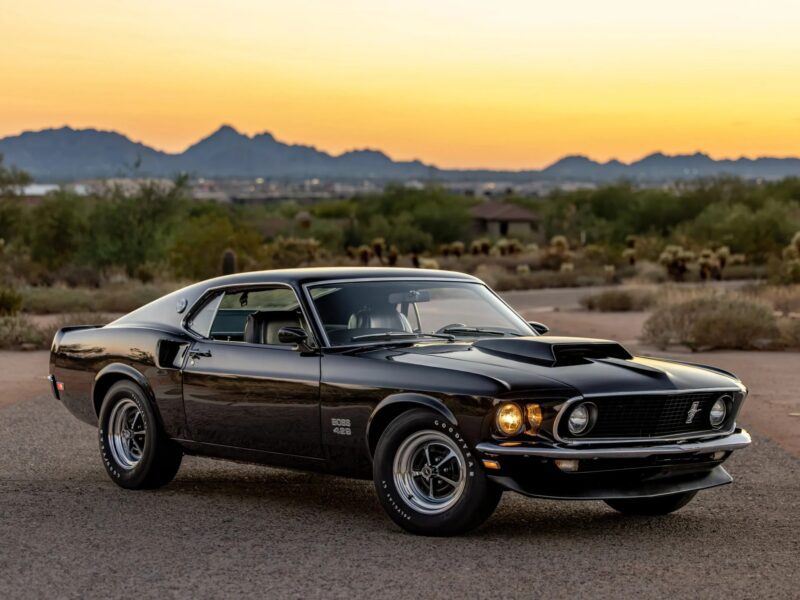 A black 1969 Ford Mustang Boss 429, a true icon among classic muscle cars, parked on a road with mountains and desert scenery in the background at sunset.
