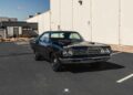 A black classic Plymouth Road Runner, a true icon among classic muscle cars, is parked in an empty lot next to a white industrial building under a clear blue sky.