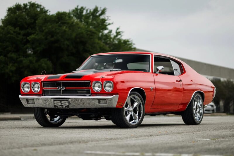 A red Chevrolet Chevelle SS 396, a true icon among classic muscle cars, is parked on pavement with trees and a building in the background.