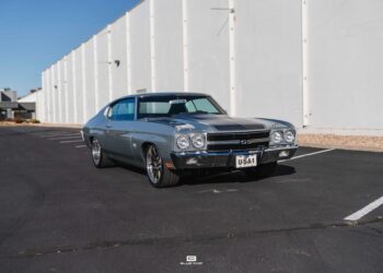 A silver 1970 Chevrolet Chevelle SS coupe, a standout among classic muscle cars, is parked on an empty asphalt lot beside a white industrial building.